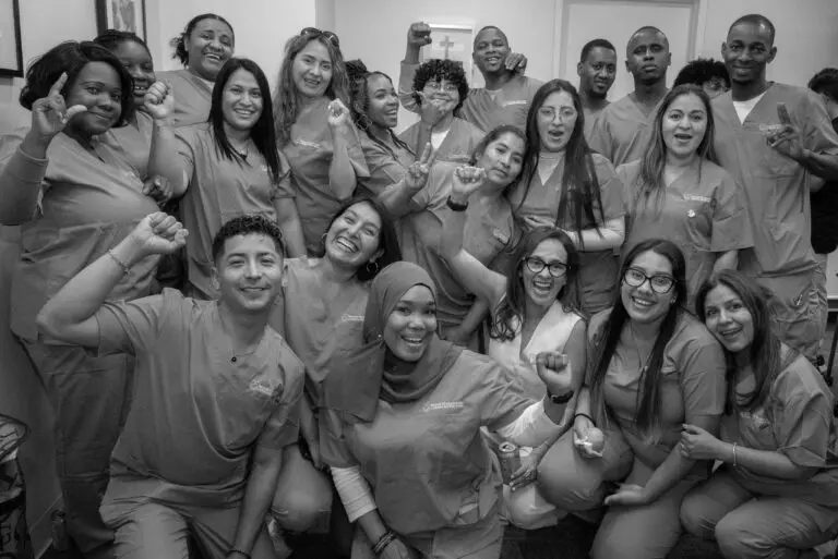 Black-and-white photo of smiling healthcare workers. They're crowded together in matching scrubs with raised fists.